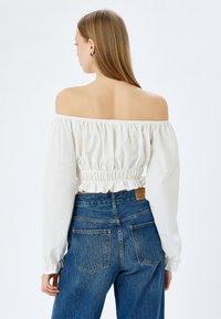 Hvid off-shoulder bluse med lange, samlede �ærmer og flæser forneden, parret med højtaljede blå denim jeans med baglommer.