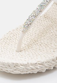 White woven flip-flop, featuring a textured braided sole, with a smooth white thong strap embellished with clear rhinestones.