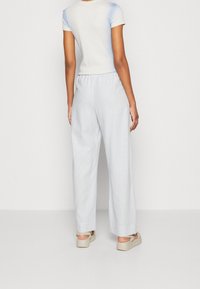 Vrouw gekleed in een lichtblauwe korte mouwen top, bijpassende loszittende broek en beige platform sandalen, gezien vanuit de rug tegen een effen achtergrond.