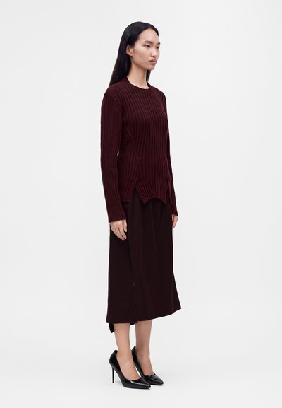 Femme debout portant un pull côtelé bordeaux foncé, une jupe midi assortie et des chaussures noires à talons hauts pointues sur un fond uni.