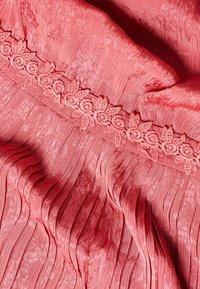 Korálová látka s texturovaným povrchem, se svislými sklady a květinovou krajkovou lemovkou podél horního okraje.