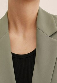 Gros plan sur le cou et le haut de la poitrine d'une personne portant un blazer vert clair par-dessus un haut noir côtelé.