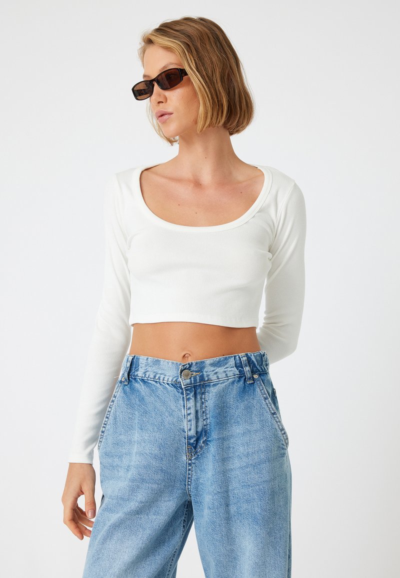 Weißes, geripptes, langärmeliges Crop-Top mit weitem Rundhalsausschnitt, kombiniert mit hochgeschnittenen, hellblauen Denim-Jeans.