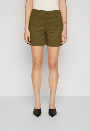 Kvinde iført olivengrønne shorts, hvid top og sorte spidse sko, der står mod en ensfarvet lys baggrund.
