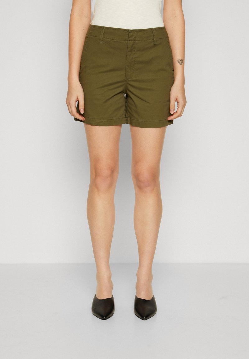 Femme portant un short vert olive, un haut blanc et des chaussures noires à bout pointu, debout devant un fond clair uni.