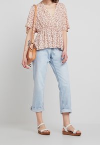 Kukapaulu floral vaaleanpunaisella kuviolla ja syvällä V-pääntiellä, yhdistettynä vaaleansinisiin, käännettyihin farkkuihin. Malli käyttää valkoisia sandaaleja ja pitää vaaleanruskeaa käsilaukkua.