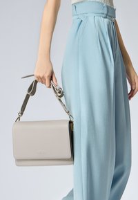 Sac à main en cuir gris avec une finition texturée et une bandoulière ajustable, tenu dans une main, aux côtés d'un pantalon taille haute bleu clair.