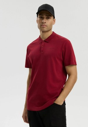 Mężczyzna w czerwonej koszulce polo i czarnej czapce, stojący z jedną ręką w kieszeni na jednolitym jasnym tle.