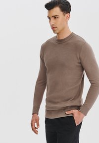 Brązowy sweter dzianinowy z teksturowanym wzorem, okrągłym dekoltem i długimi rękawami, w połączeniu z czarnymi spodniami. Gładka tkanina i dopasowany krój.