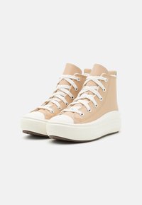 Zapatillas altas beige hechas de cuero suave, con cordones blancos, puntera de goma blanca y una suela gruesa y texturizada.