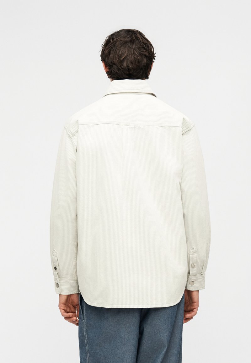 Mann mit dunklen Haaren, der ein hellbeiges Cordhemd und lockere blaue Hosen trägt, steht mit dem Rücken zur Kamera vor einem schlichten Hintergrund.