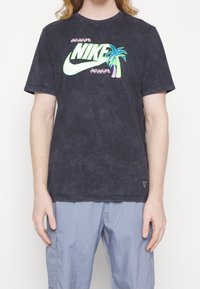 Černé tričko s vázáním Nike má barevnou grafiku palmy a loga. Kulatý výstřih, krátké rukávy, měkká tkanina a volný střih.