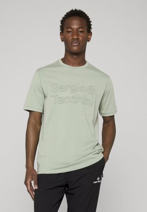 Uomo in piedi che indossa una maglietta verde chiaro Sergio Tacchini e pantaloni neri con il logo Sergio Tacchini, su sfondo grigio chiaro.