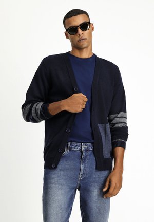Cardigan blu navy con chiusura a bottoni, maniche a righe grigie a contrasto, lavorazione a maglia testurizzata e tasca frontale. Indossato sopra una t-shirt scura.