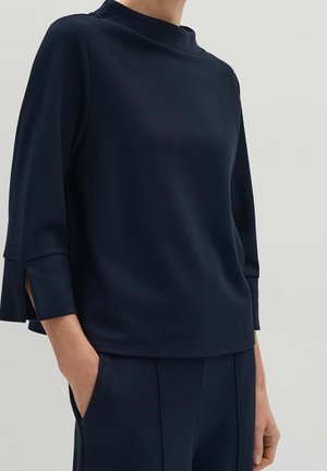 Persoon draagt een marineblauw textuur top met driekwart mouwen en zijslitten, gecombineerd met bijpassende marineblauwe broek, handen in de zakken.