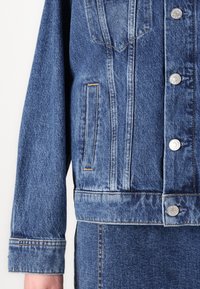 Denim jacket in medium blue. Features button-up front, two chest pockets, and stitched detailing. Textured fabric with a slightly worn appearance.