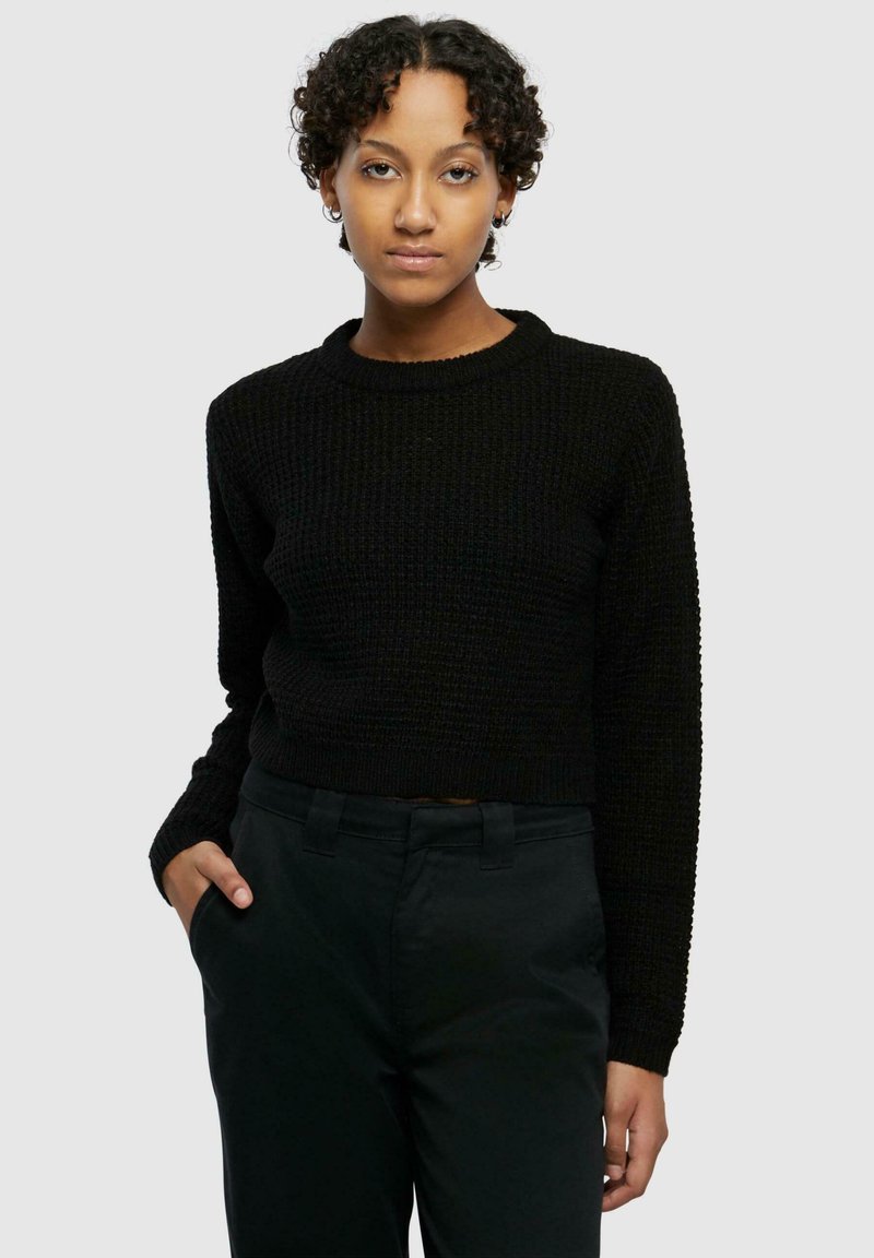 Maglione corto nero a maglia con texture a coste, scollo rotondo e maniche lunghe, abbinato a pantaloni scuri.