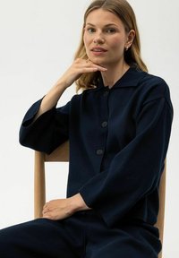 Maglione blu navy, con texture, dotato di colletto e tre bottoni, indossato con maniche rilassate. Modello seduto, mano poggiata sul mento.