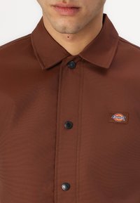 Camicia marrone con bottoni, realizzata in tessuto morbido, con colletto, bottoni neri e un piccolo patch con logo sul petto.