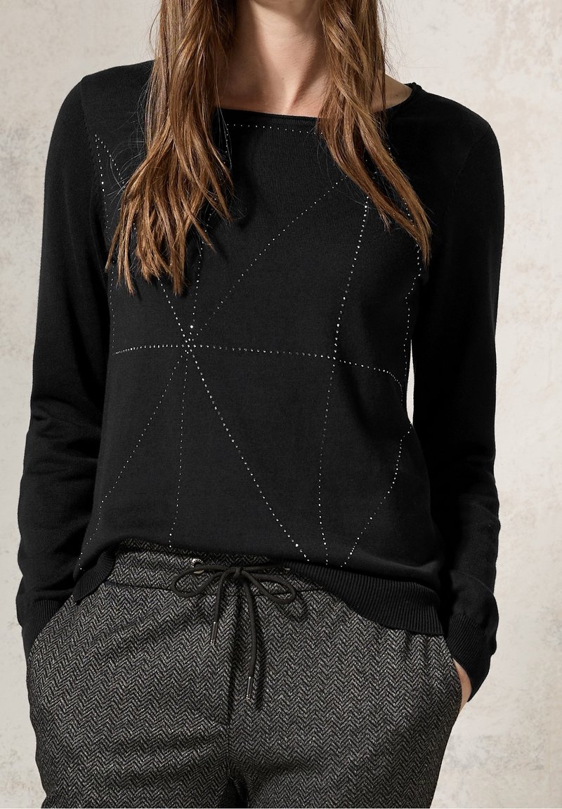 Kvinna i svart långärmad tröja med subtil prickad geometrisk design och grå herringbone byxor med dragsko, händerna i fickorna.