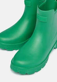 Zelené gumové členkové čižmy s matným povrchom, textúrovanými podrážkami a embosovaným logom značky na boku. Pevná, vodotesná konštrukcia pre vonkajšie použitie.