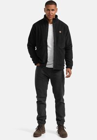 Sort fleece-jakke med lynlås foran, standkrave og to for-lynlåsede lommer. Bæret over en hvid skjorte, parret med mørke jeans og brune støvler.