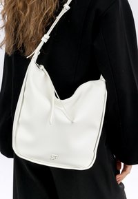 Borsa a tracolla bianca in pelle con design arrotondato, texture liscia e chiusura con zip. Decorata con un piccolo logo impresso sul davanti.