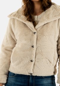 Chaqueta de piel sintética de color crema con cuello de chal, cinco botones grises y una textura suave, combinada con un suéter ligero por debajo.