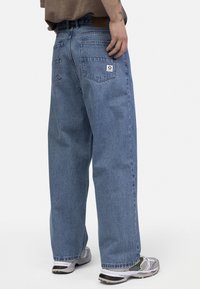 Blugi din denim albastru deschis cu o croială relaxată, având două buzunare la spate și o etichetă din piele pe talie, purtați cu pantofi sport gri.