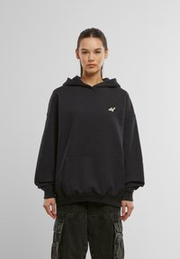 Czarnoszary, oversize hoodie wykonany z miękkiego materiału, z przednią kieszenią i małym beżowym logo na klatce piersiowej. W zestawie z kapturem ze sznurkiem.