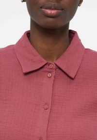 Κλείσιμο σε ένα άτομο που φοράει πουκάμισο με υφή σε χρώμα σκονισμένο ροζ, με μύτη γιακά και μικρά στρογγυλά κουμπιά.