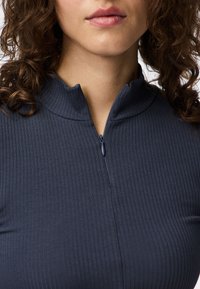 Mulher vestindo uma blusa de malha azul-marinho com um fecho parcialmente fechado perto do pescoço, cabelo castanho encaracolado visível nos ombros.