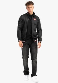 Zwarte zip-up jas met witte en rode gestreepte accenten, gecombineerd met een zwart T-shirt en donkere jeans. Model draagt witte sneakers.