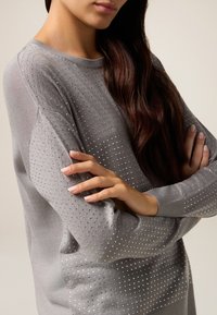 Grau gestrickter Pullover mit strukturiertem Muster und kleinen Löchern, gerippten Bündchen und einer lockeren Passform. Akzente in Form weißer Punkte fügen Details hinzu.