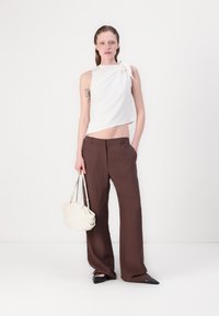 Hvit ermeløs topp med knute detalj, kombinert med mørkebrun vide bukser. Modellen holder en lys beige håndveske og har på seg svarte spisse sko.