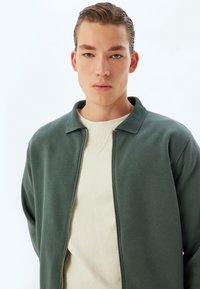 Chaqueta verde con cremallera y cuello acanalado sobre un suéter beige claro. Las telas parecen suaves con una textura lisa y un diseño casual.