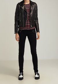 Czarna skórzana kurtka motocyklowa z srebrnymi suwakami, sweter w czerwono-czarny wzór, wąskie czarne dżinsy i czarno-białe buty.