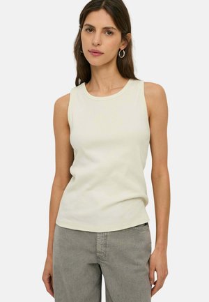 Mujer con cabello largo y castaño, que lleva pendientes de aro plateados, una camiseta blanca sin mangas con textura acanalada, y pantalones grises sobre un fondo liso.