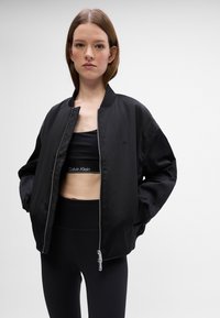 Fekete bomber dzseki sima textúrával és bordázott mandzsettával, cipzáros záródással és diszkrét logóval. Fekete cropped top-pal és leggingsekkel párosítva.