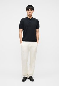 Άνδρας που φοράει μαύρο πόλο μπλουζάκι με κοντά μανίκια, λευκό φαρδύ παντελόνι και μαύρα λουστρίνια, στέκεται με τα χέρια στις τσέπες μπροστά σε απλό φόντο.