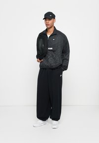 Svart nylon vindjakke med mønster, med en frontlomme og "AIR" tekst, paret med løse svarte bukser og hvite sneakers.