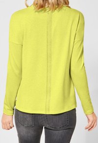 Långärmad skjorta i klargul, med en ryggsöm som har en glänsande detalj. Mjukt tyg med en avslappnad passform, ihop med mörka jeans.