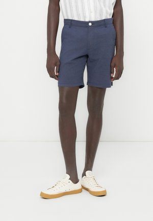 Donkerblauwe korte broek van een lichtgewicht stof, voorzien van een platte voorkant, knoopsluiting en omgeslagen zoom, gecombineerd met beige sneakers.