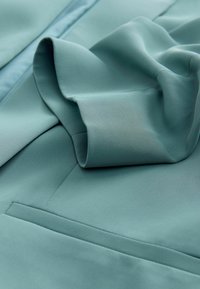 Tissu bleu-vert clair plié lisse avec des coutures visibles et un petit détail de poche sur une surface textile douce.