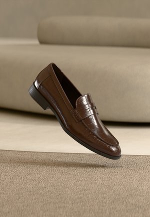 Mocassin en cuir marron avec un motif texturé flottant au-dessus d'un tapis beige avec un canapé beige assorti en arrière-plan.