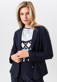 Navy blazer in een slim fit, met een geknipte revers en twee knopen. Het model draagt een blouse met een geometrisch patroon eronder.