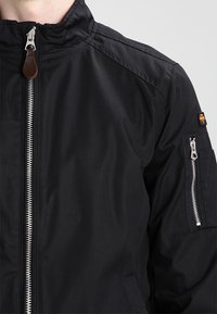 Giacca nera con zip frontale, colletto alto e tasche con zip sulle maniche. Texture liscia con dettagli di design sobri.