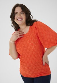 Ljus orange strukturerad topp med ett diamantmönster, vida ärmar och rundad halsringning, kombinerad med mörkblå jeans.