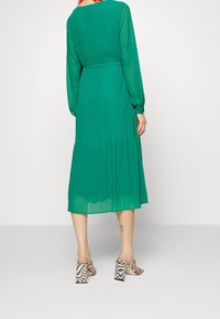 Zelené šaty s dlouhým rukávem, s plisovanou sukní a přiléhavým pasem. Modelka má na sobě kostkované sandály na vysokém podpatku, které ukazují hladkou a splývavou tkaninu.