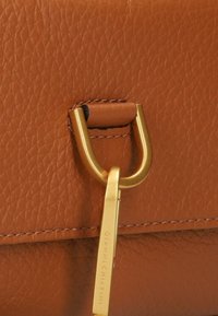 Detail van een bruine leren handtas met een goudkleurige metalen gesp en een sleutelhanger met gegraveerde merknaam. Geweven oppervlak met nette stitching.